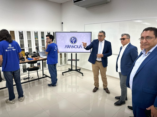 Foto mostra homens reunidos numa sala, com um telão ao fundo