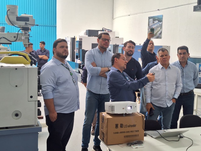 Foto mostra homem vestido de blusa azul e usando óculos mostrando uma máquina a grupo de pessoas