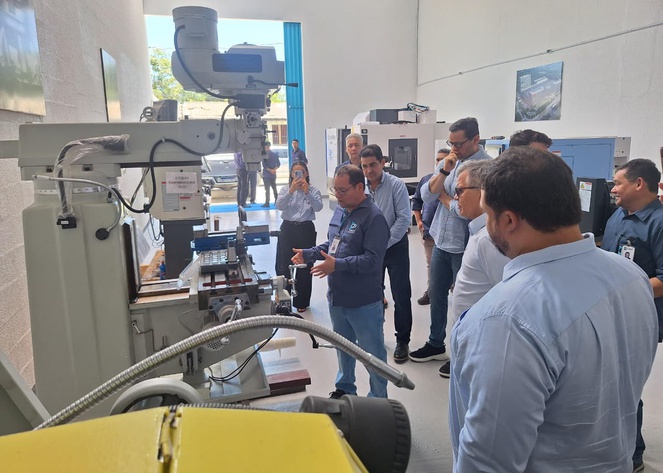 Foto mostra um homem de blusa azul e óculos mostrando uma máquina a grupo de pessoas