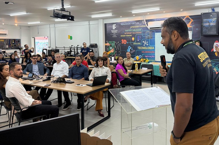 Foto mostra homem barbado, de blusa preta e microfone na mão falando em um evento