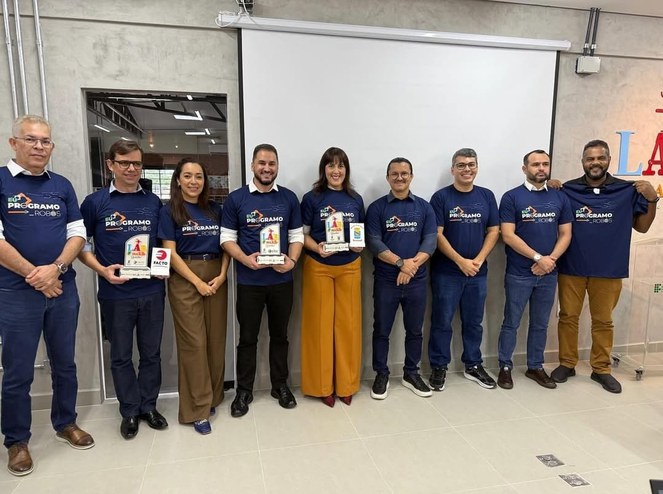 Foto mostra um grupo de pessoas vestidas com blusa azul e mostrando troféus nas mãos
