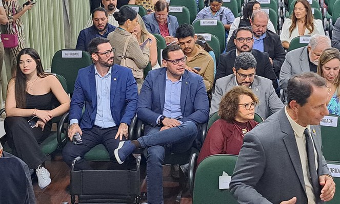 Foto mostra dois homens de blazer azul e óculos sentados em um auditório