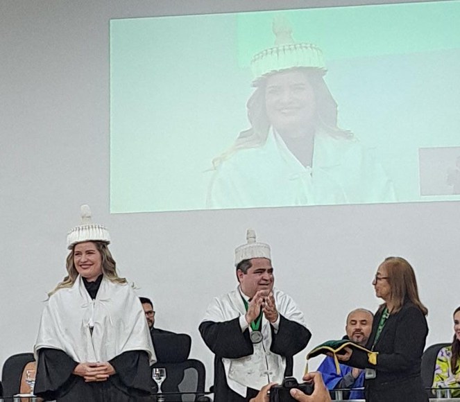 Foto mostra mulher com bata e chapéu brancos recebendo uma homenagem