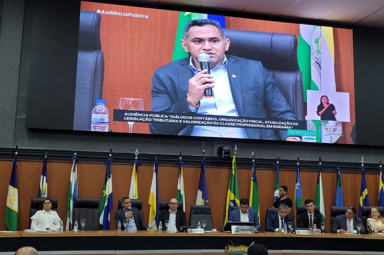 Foto mostra homem de paletó azul falando em um evento público e mostrado também num telão