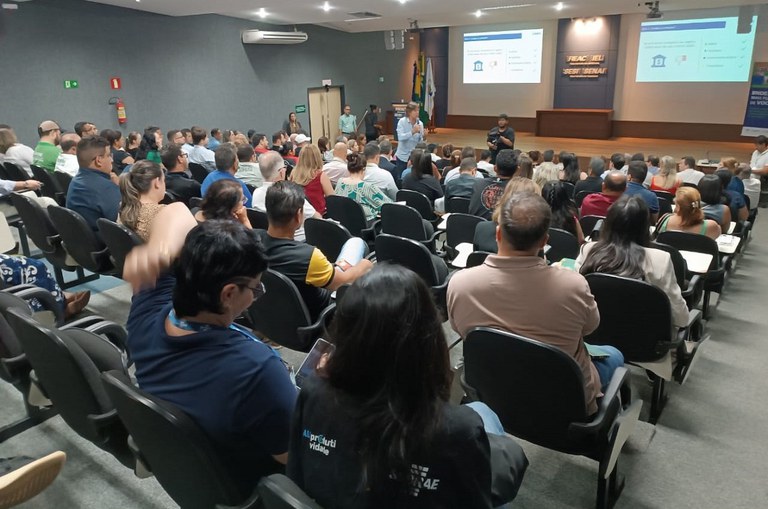 Foto mostra homem em pé com microfone na mão falando em público