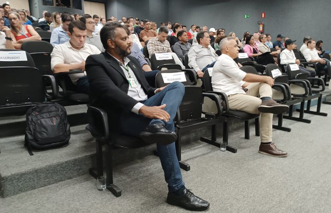 Foto mostra homem de barba, paletó preto e calça azul observando uma explanação em evento