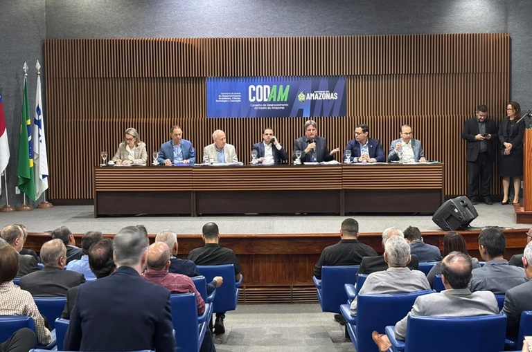 Foto mostra homem falando ao microfone à mesa, num evento realizado num auditório lotado e com um telão ao fundo