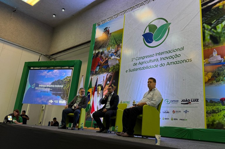 Foto mostra três homens sentados um ao lado do outro, com o do meio utilizando o microfone para falar ao público presente no evento