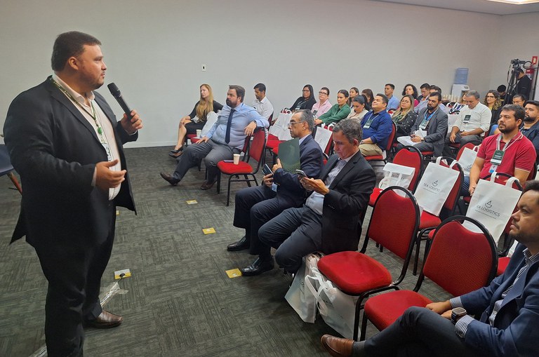 Foto mostra homem de blazer preto e calça preta, ministrando palestra em um evento
