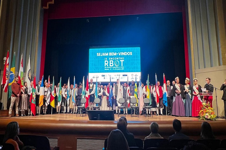 Foto mostra grupo de pessoas segurando bandeiras e uma mulher de preto falando ao microfone