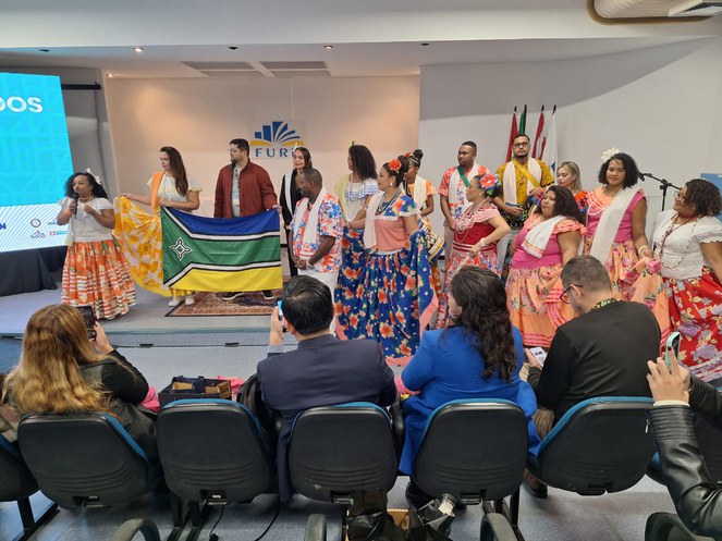 Foto mostra mulher vestida com saia colorida falando a um grupo de pessoas