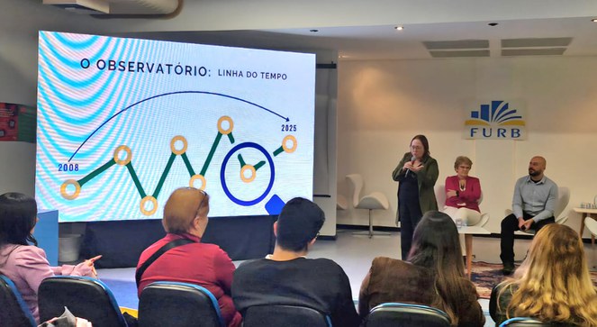 Foto mostra mulher em pé com microfone na mão fazendo uma explanação com um painel grande como apoio