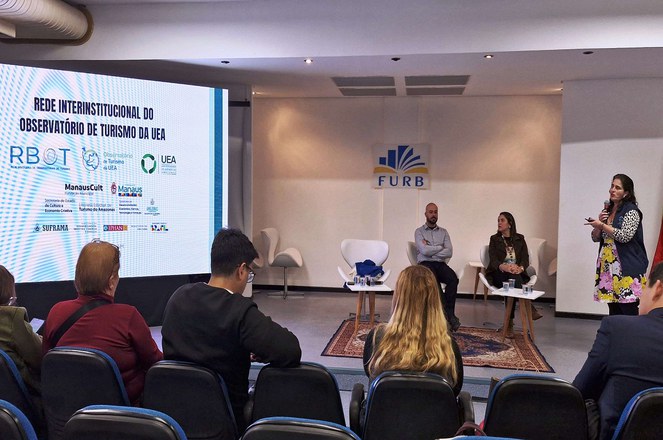 Foto mostra mulher em pé fazendo uma explanação com o apoio de informações no telão