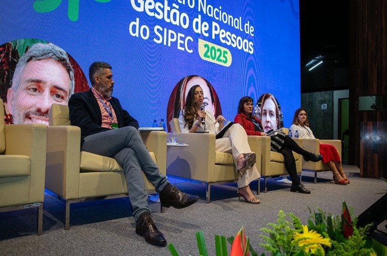 Foto mostra mulher falando ao microfone, observada por outras pessoas