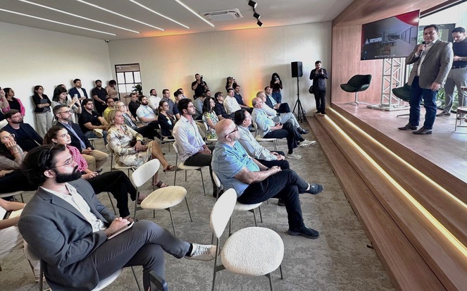 Foto mostra um homem de blazer cinza, microfone na mão e um telão ao fundo, falando em evento público