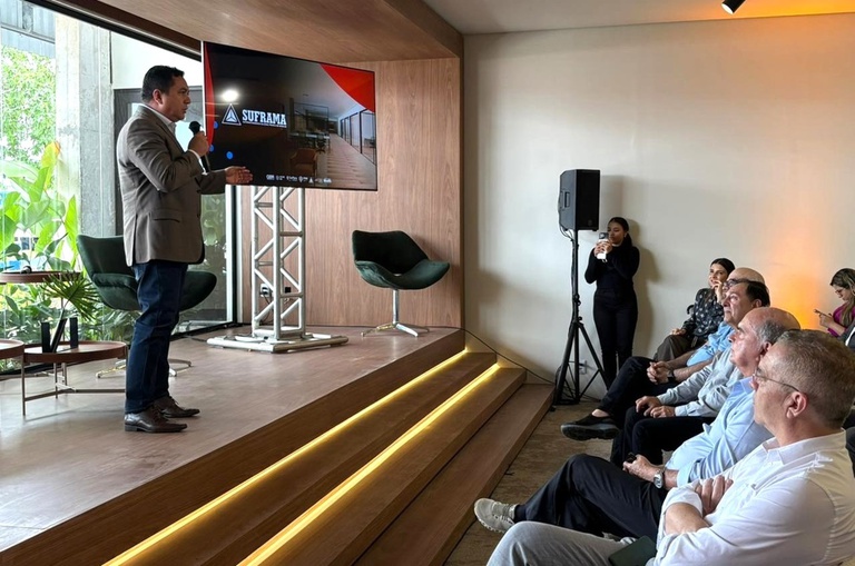 Foto mostra homem de blazer cinza, microfone na mão e um telão ao fundo, falando em evento público