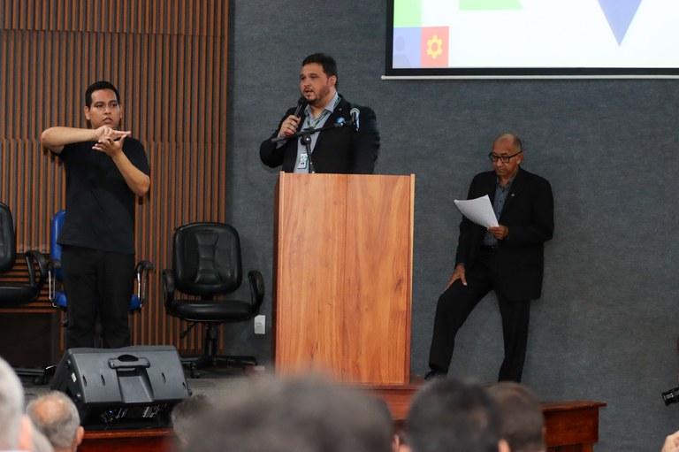 Foto mostra homem de blazer escuro e camisa azul, falando com microfone na mão