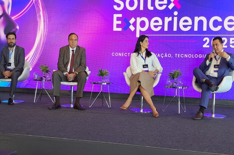 Foto mostra homem falando ao microfone, observado por uma mulher e outros dois homens