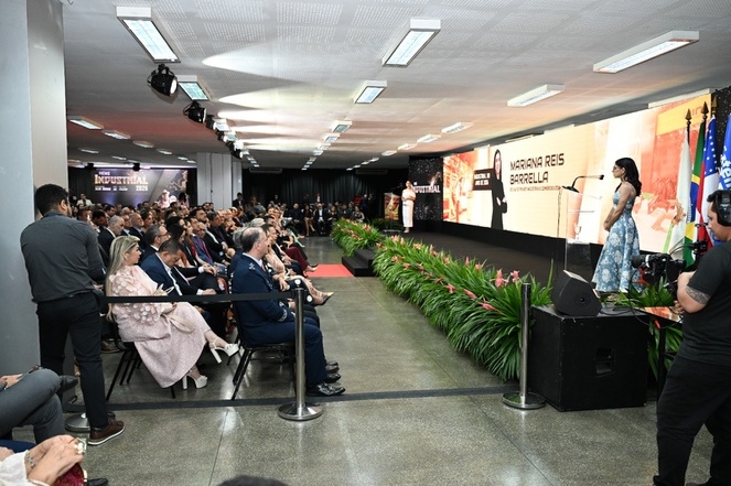 Foto mostra mulher em pé, observas por pessoas sentadas num evento