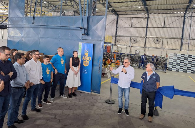 Foto mostra homem falando ao microfone, em preparação para o descerrando de uma fita de inauguração numa fábrica