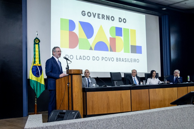 Foto mostra homem em pé de blazer falando ao microfone, observado por grupo de pessoas e com um telão ao fundo