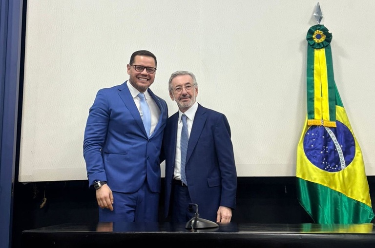 Foto mostra dois homens de blazer posando para foto ao lado de uma bandeira