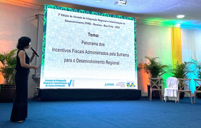 Foto mostra uma mulher com microfone na mão proferindo uma palestra com o auxílio de um telão ao fundo