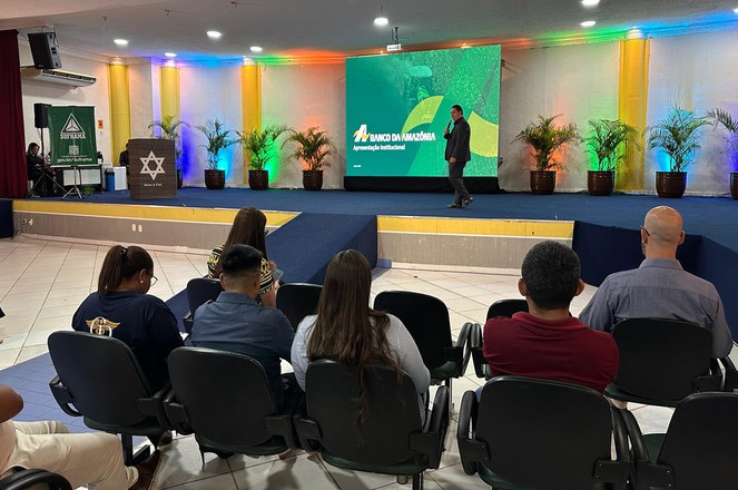 Foto mostra homem com microfone na mão proferindo uma palestra com o auxílio de um telão ao fundo