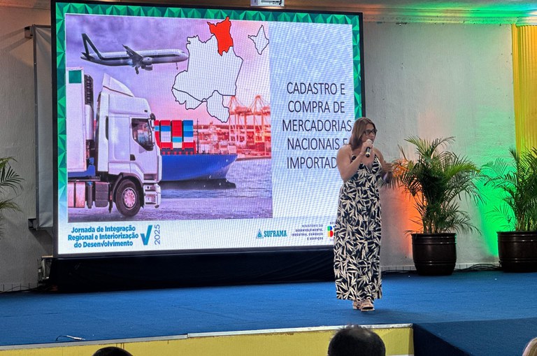 Foto mostra mulher com microfone na mão proferindo uma palestra com o auxílio de um telão ao fundo