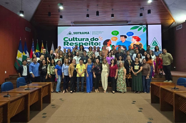 Foto mostra mulher grupo de pessoas em pé posando para sessão de foto, com um telão ao fundo
