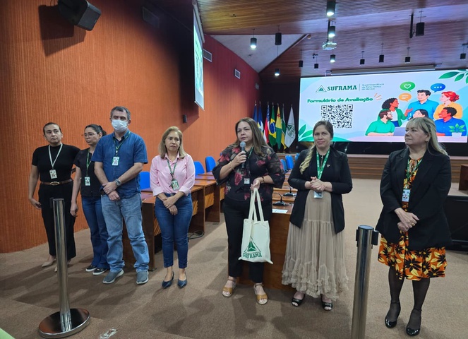 Foto mostra pessoas em pé e uma delas falando ao microfone, com um telão ao fundo