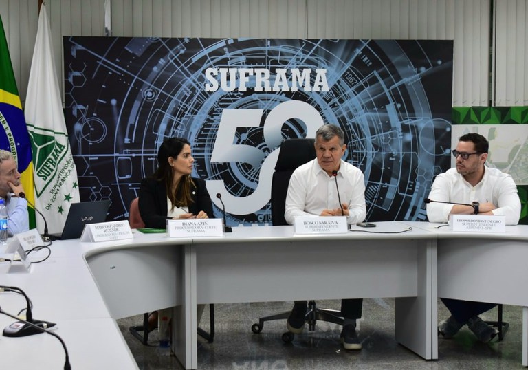 Foto mostra homem de blusa branca falando, ao lado de outro homem que também está de blusa branca além de óculos, e de uma mulher com blazer preto