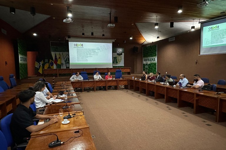 Imagem mostra pessoas sentadas em um auditório com mesa em U