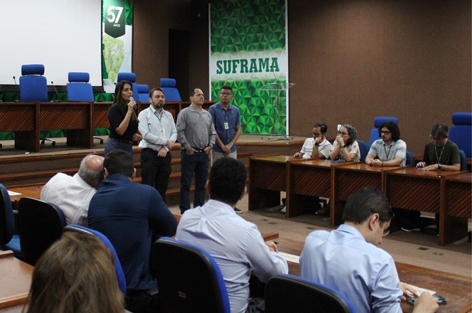 Foto mostra mulher falando ao microfone, observada por grupo de pessoas