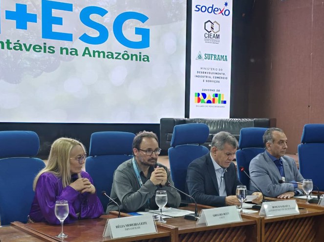 Foto mostra homem falando ao microfone, ao lado de outras pessoas e tendo um telão ao fundo