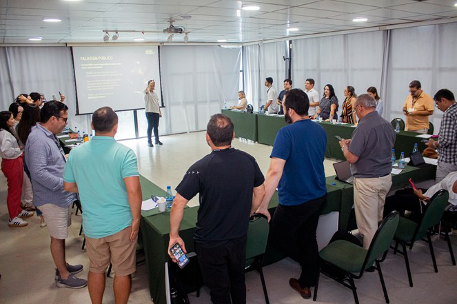 Foto mostra homem fazendo uma dinâmica com um grupo de pessoas numa sala, com o apoio de um telão