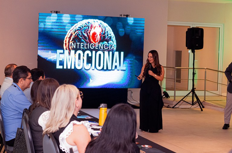 Uma mulher a frente, de pé, apresentando informações a um público sentado em fila