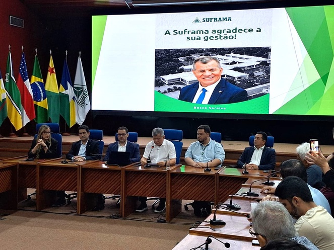 Foto mostra homem falando ao microfone, observado por grupo de pessoas e com um telão ao fundo