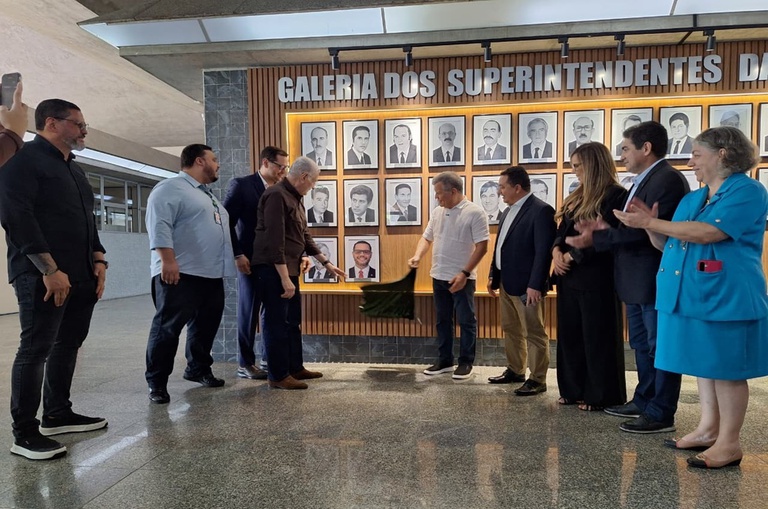 Foto mostra homem de blazer assinando um documento, com outros homens observando