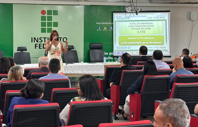 Foto mostra mulher em pé ministrando uma palestra com um telão ao fundo