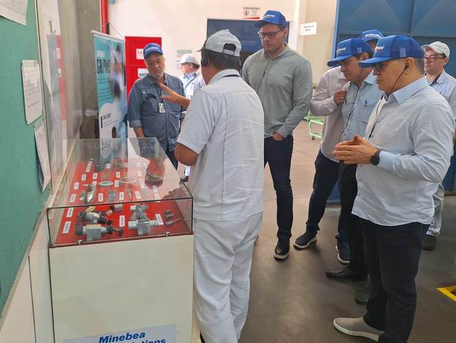 Foto mostra homem mostrando exposição de produtos a outros homens, durante visita técnica a uma fábrica