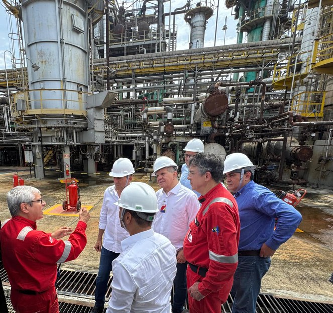 Foto mostra homem de macacão vermelho fazendo uma explicação a grupo de homens sobre o funcionamento de maquinárias ao fundo