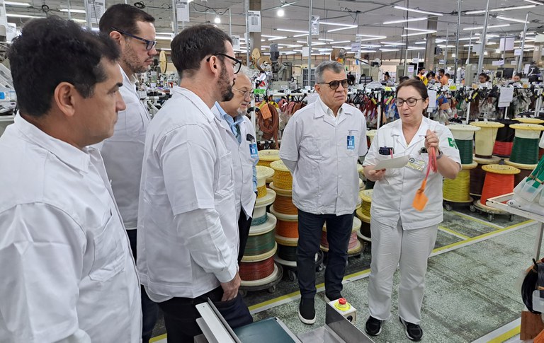 Foto mostra uma mulher de bata branca com papel e um cabo na mão passando informações a um grupo de pessoas