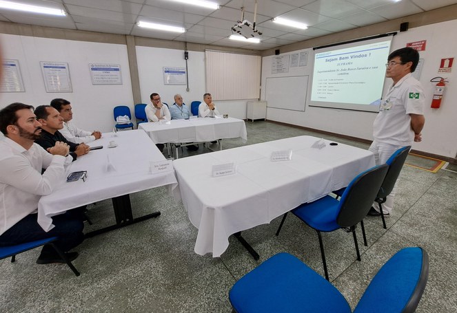 Foto mostra homem em pé e de óculos fazendo uma explanação com um telão ao fundo