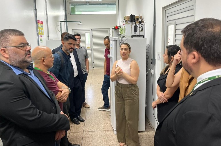 Foto mostra mulher de blusa bege e calça marrom fazendo uma explicação a grupo de pessoas em um corredor