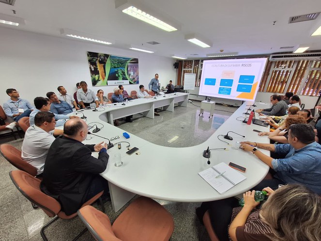 Foto mostra pessoas em reunião numa sala com o apoio de um telão