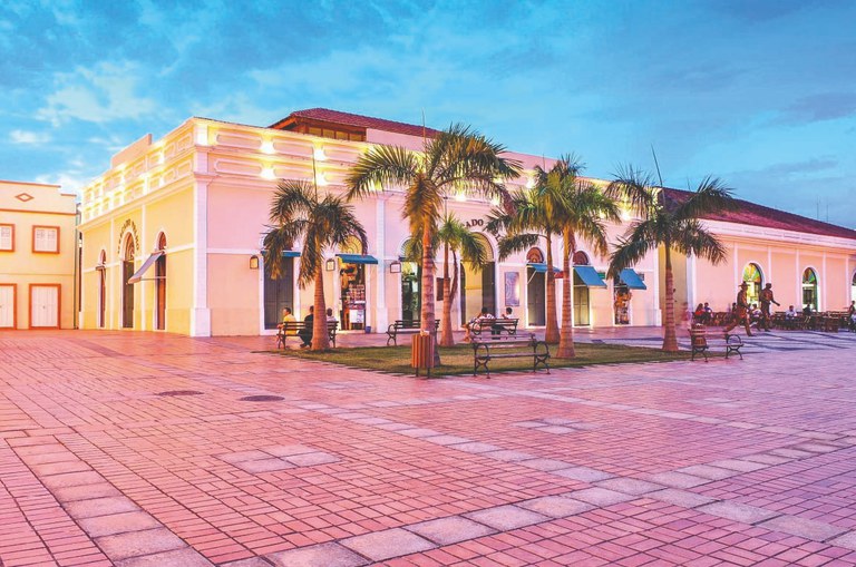 Foto mostra uma praça ampla com piso de ladrilhos rosados e palmeiras alinhadas diante de um prédio histórico iluminado, de fachada clara e janelas arqueadas ao entardecer.
