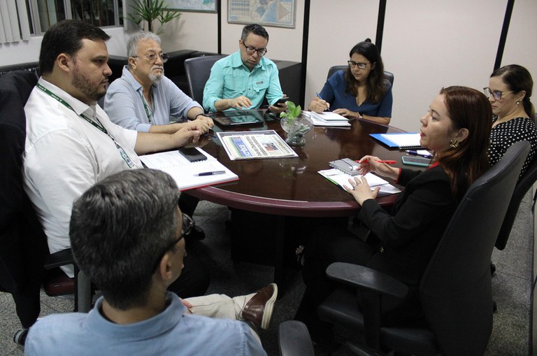 Foto mostra pessoas reunidas em uma sala fechada, com jornal e cadernos de anotação à mesa
