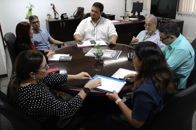 Foto mostra pessoas reunidas em uma sala, numa mesa com jornal e caderno de anotações