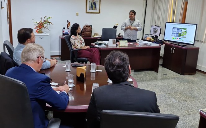 Foto mostra homem em pé fazendo uma explanação a grupo de pessoas com o apoio de um televisor